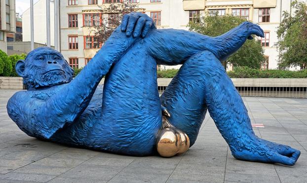 A bronze sculpture titled "King Kong Balls" by Denis Defrancesco, depicting a blue gorilla lying on its back with its legs crossed, showcasing exaggerated golden testicles. The artwork, measuring 5.04 meters long, 2.10 meters wide, and 2.25 meters high, and weighing 2780 kg (6128 lbs.), is placed in an urban plaza with modern buildings and greenery in the background. The vivid blue of the gorilla contrasts strikingly with the metallic gold elements, creating a bold visual statement.