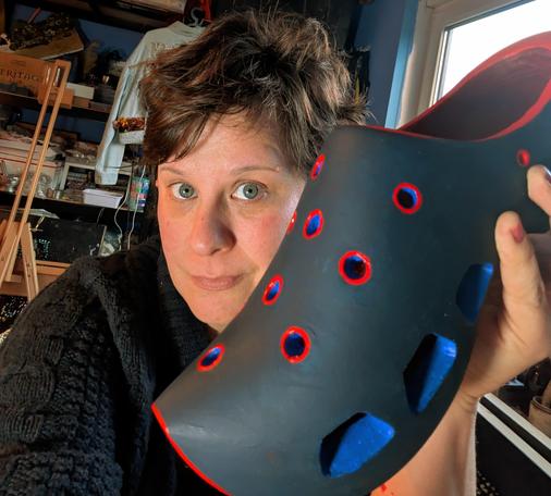 A middle aged white woman with a brunette pixie cut sitting in an art studio. She is holding a clog up to the camera. It's been carved into o resemble a Croc. It's dark blue. The holes are painted light blue and rimmed in red.