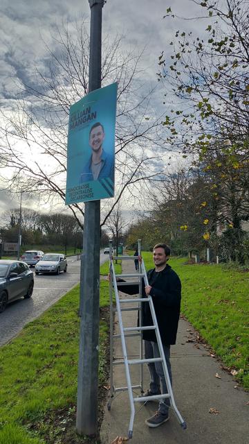 A photo of Ciarán smiling while holding the ladder beside a poster.