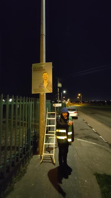 A photo of James pointing at something in the distance while standing beside a ladder and poster.
