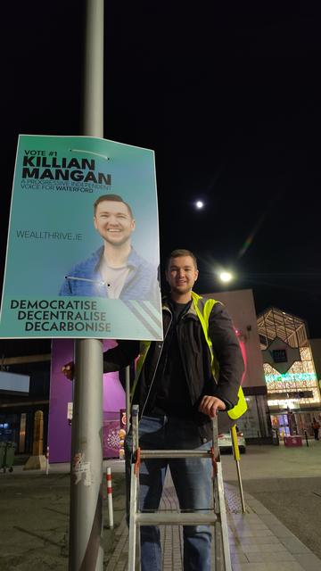 A photo of Killian smiling and standing on a ladder beside a poster.