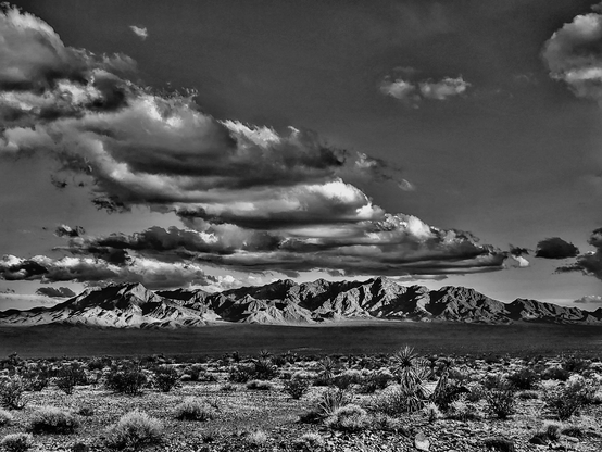 In the remote corners of the desert the sun shines on a lonely mountain range while clouds wisp above laying a shadow across the plains.