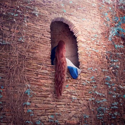 Girl with blue dress and long brown hair sits in arched stone castle window