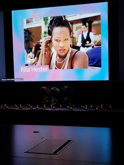 A dark room with a screen that has the words Rita Hester on it, along with an image of her. Below are numerous electric tea candles on top of a trans flag
