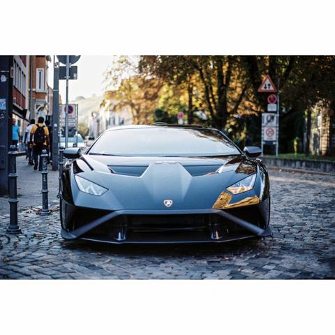 The pictures shows the full front of a black lamborghini in an old street of Esslingen, with trees and signs in the background.
