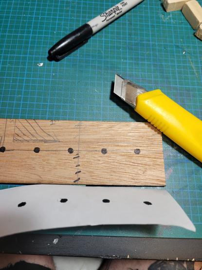 A piece of wood with a paper template to mark drill holes. A sharpie was used to mark the places to drill and a box cutter lays beside the piece of wood