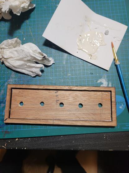 After holes were drilled into the piece of wood another wooden frame is glued on top to frame the 5 holes. Paper towel, and a large glob of wood glue and a paint brush sit above