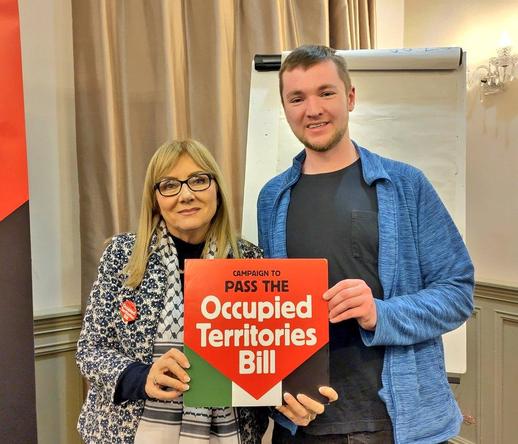 A photo of Senator Frances Black and Killian holding up a sign reading "Campaign to Pass the Occupied Territories Bill" in the colours of the Palestinian flag.
