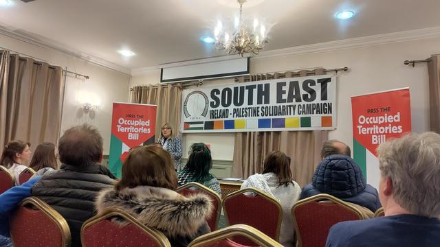 A photo of Senator Frances Black speaking to a crowd with a sign reading "pass the occupied territories bill" and another sign reading "South East Ireland - Palestinian Solidarity Campaign" behind here.