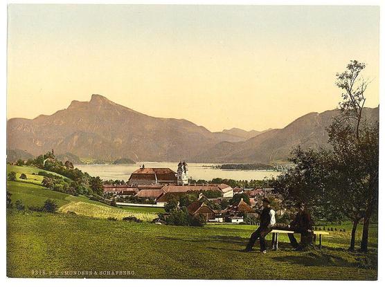 The image depicts a serene landscape scene, likely from around the late 19th century based on its style. In the foreground, two individuals are seated at a picnic table in a grassy area with trees scattered around them. They appear to be engaged in conversation or relaxation.

In the midground, we see an expansive view of rolling hills leading up to distant mountains that dominate the background. Nestled amidst these green expanses is a large village or town characterized by red-roofed buildings and what appears to be a church with prominent spires. The architecture suggests European influences from around this time period.

A calm lake reflects the surrounding scenery, adding tranquility to the overall composition. A notable feature of the image is its coloration; it has been printed in sepia tones which were commonly used for photographic prints during that era. This gives the scene a warm, nostalgic quality.

The caption below indicates "Mondsee & Schafberg," suggesting these are key locations depicted within this picturesque view. Mondsee and Schafberg refer to specific landmarks or regions; Mondsee being a lake in Upper Austria known for its natural beauty, while Schafberg is likely referring to the nearby mountain range where one might find such leisurely pastimes as seen here.

Overall, the image captures an idyllic moment of leisure and enjoyment amidst nature's splendor within what was possibly a resort area dur [...]