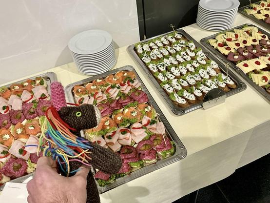 Max, das bunte Strickhorn, schaut gierig auf mehrere große Tabletts mit verschiedenen Snacks.