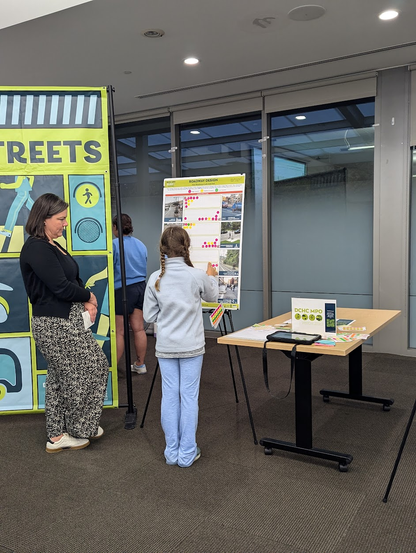 Photo of a woman standing next to a pre-teen girl, both looking at a poster. The poster is posing questions about people's concerns about street design. The girl is putting stickers on the poster.