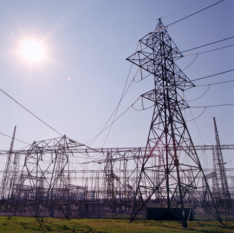 The sun forms a star in the upper left and lens flares radiate diagonally down to the right over an extensive transformer field. A pylon stands tall on the right side of the frame.