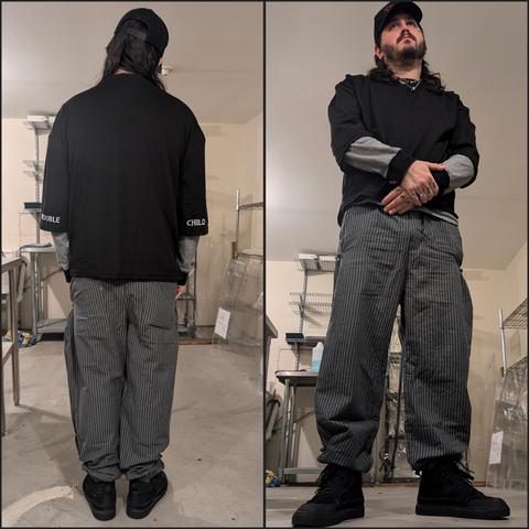 A person is shown in a collage of two images wearing a Japanese streetwear-inspired outfit. In the left image, the back of the outfit is visible, featuring an oversized black shirt with text on the sleeves ("TROUBLE" on the left sleeve and "CHILD" on the right sleeve), paired with grey and white vertically striped harem-style pants. The person is also wearing a black cap and black shoes. In the right image, a front view shows the person standing with their arms crossed, highlighting the layering of a long-sleeve shirt with thin black and white stripes underneath the oversized black shirt. The relaxed fit of the pants and monochromatic tones emphasize a casual, streetwear aesthetic. The setting appears to be a minimalist workshop or storage space with metal shelves and a concrete floor.
