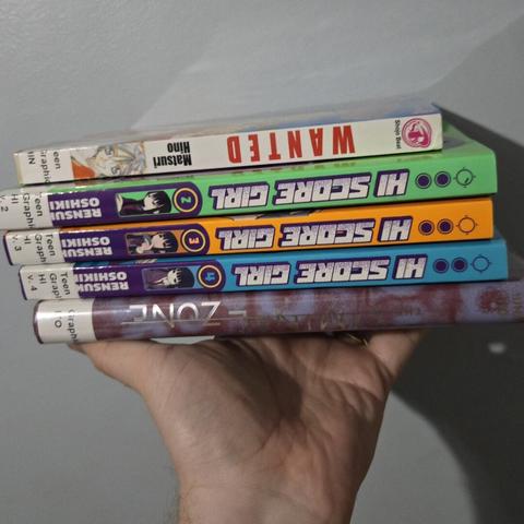 Photograph of a white person's hand holding a stack of books against a light colored wall. The spines are visible. From top to bottom, the book spines read: Wanted, Hi Score Girl 2, Hi Score Girl 3, Hi Score Girl 4, The Liminal Zone.