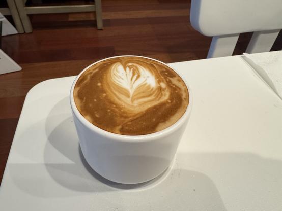 Small white coffee mug filled with a cappuccino containing dark and light brown mixed with white latte patterns sitting on a white table with dark brown hardwood floor in the background.