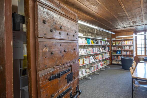 Inside of a former prison cell with stone walls and a timber roof. A heavy wooden door with iron fittings and a peephole is propped open to the left of the image. Converted into a library, the room has shelves along the walls and a table in the middle. A barred window pierces the back wall.