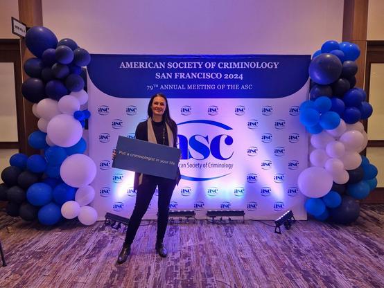 Das Bild zeigt Dr. Nicole Selzer von der Cyberagentur bei der American Society of Criminology (ASC) 2024 in San Francisco. Sie steht vor einer Fotowand mit dem Logo der ASC, umgeben von blau-weißen Luftballons. In ihren Händen hält sie ein Schild mit der Aufschrift „Put a criminologist in your life“. Die Szene ist hell und freundlich beleuchtet, und Dr. Selzer lächelt in die Kamera. Das Foto vermittelt die professionelle, aber auch einladende Atmosphäre der Veranstaltung. 
Foto: privat