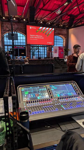 Das Bild zeigt eine Indoor-Umgebung mit einem großen digitalen Mischpult im Vordergrund, was auf eine Toningenieur- oder Live-Event-Produktionsumgebung hinweist. Das Mischpult hat zahlreiche Knöpfe, Tasten und Bildschirme, die auf komplexe Audiosteuerungsmöglichkeiten hinweisen. Im Hintergrund sitzt ein Publikum, das von der Kamera weg auf eine große Präsentationsleinwand blickt. Auf der Leinwand steht: "Herzlich willkommen! Schön, dass Sie da sind." Der Veranstaltungsort scheint eine große Halle mit hohen Decken und freiliegenden Backsteinwänden zu sein, typisch für industrielle oder Loft-ähnliche Räume, die oft für Veranstaltungen oder Konferenzen genutzt werden.