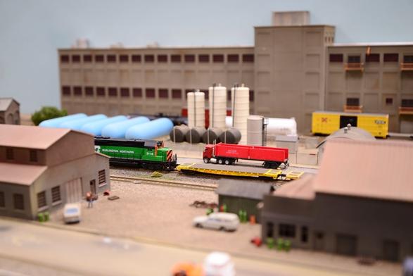 An N-scale Burlington Northern locomotive pulls some flatcars through an industrial area.