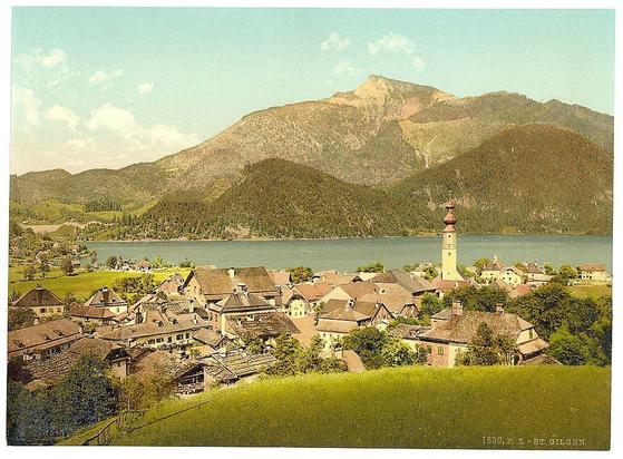 The image depicts a picturesque village nestled amidst lush green hills and mountains. It appears to be from the late 19th or early 20th century, as suggested by its vintage style.

In the foreground, there's a large expanse of vibrant green grass, leading up to what seems like a gently sloping hillside that extends into the background where quaint houses with steeply pitched roofs are scattered. These structures likely represent traditional architecture common in rural Austrian villages during this period.

The village is set beside a calm lake or river reflecting clear blue skies and surrounding foliage. The water adds a sense of tranquility to the scene, enhancing its idyllic charm.

Dominating the central part of the image is an imposing church tower with spires reaching upwards toward the sky, possibly indicating it as a significant landmark within this community. This structure stands out against the natural backdrop due to both its height and distinctive architectural style.

The background features rolling hills adorned with dense forests in varying shades of green, suggesting rich biodiversity typical for such European landscapes during late spring or early summer seasons. The distant mountains are less defined but still contribute significantly to the overall picturesque quality of this scene.

In summary, it's a postcard-like illustration capturing an idyllic village setting within rural Austria at around 1890 [...]