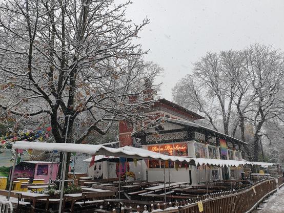 Le café Rosa Bonheur aux Buttes-Chaumont sous la neige. Les néons de l'enseigne ressortent dans la luminosité faible; la terrasse est vide et pleine de neige; il y a des arbres dont les branches sont nues et couvertes de neige; le ciel est blanc.