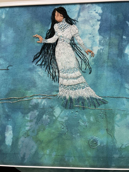 An embroidered picture of La Llorona standing partially submerged in the Rio Grande with outlines of faces and reaching hands in the water below her