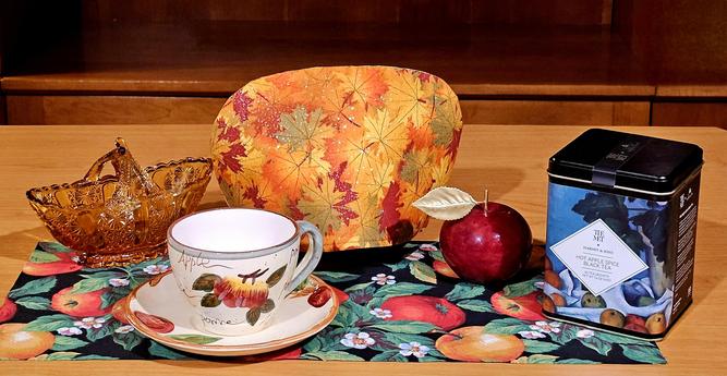 A tea tableau of fabrics in Autumn leaf and apple patterns. There is vintage china with apple motifs, an amber Depression glass basket, a red marble apple from Volterra, Italy, and Hot Apple Spice black tea from Harney and Sons.