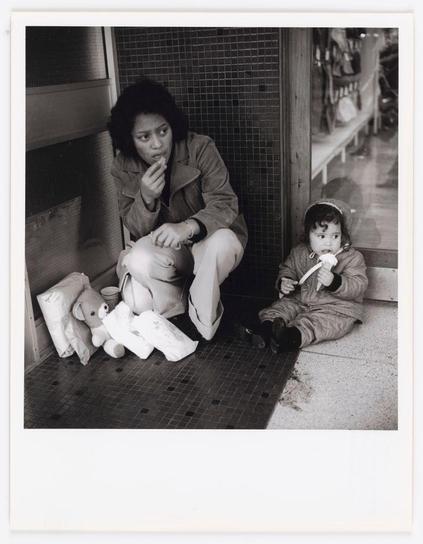 The image is a black and white photograph depicting two individuals sitting against a tiled wall on the floor. To the left, there's an adult with dark hair, wearing what appears to be a jacket or coat over casual clothing. They are holding their chin thoughtfully in one hand while resting it on their arm. Their gaze seems directed off-camera.

On the right side of the image is a child, dressed in light-colored pants and a long-sleeved top, possibly a hooded garment. The child appears to be sitting with legs outstretched straight ahead or slightly towards themselves, holding what looks like a bottle or cup near their mouth with both hands. In front of them are several large, white items that resemble oversized pillows.

Beside the individuals on the floor is an assortment of objects including teddy bears and other plush toys, which suggests they might be homeless or living in temporary circumstances. The overall mood conveyed by this photograph evokes feelings of hardship and contemplation amidst a seemingly everyday setting.