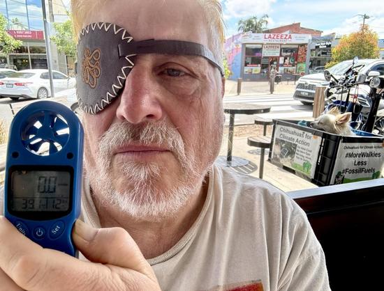 A person with a pirate eye patch sitting at an outside cafe holds up an anonemter showing the temperature. Two chihuahuas sit in a bike basket in the background.