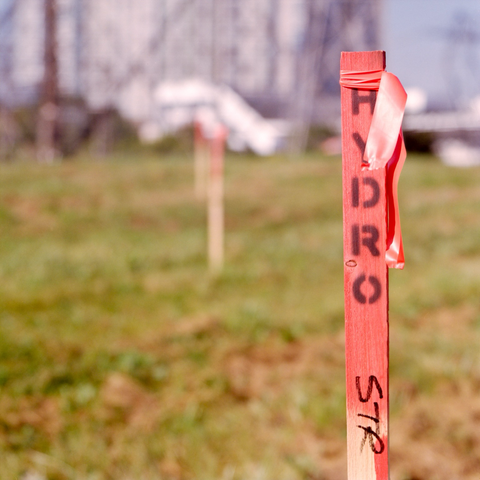 A wooden stake, painted red and topped with a red ribbon, stenciled in black “HYDRO” vertically from the top, stands in clear, shallow focus against a green field, other similar stakes blurry in the distance.