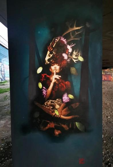 Streetartwall. A colorful mural was sprayed/painted on a round, grey concrete pillar under a road bridge. The background is dark green with dark tree trunks. A forest woman appears in the middle of this forest. The beautiful woman sits cross-legged between leaves and branches and asks us to be quiet with a finger on her lips. She is wrapped in fur and wears a voluminous headdress made of flower-decorated deer antlers. Small green leaves swirl through the air. An enchanting creature with a somewhat strange taste in clothes.