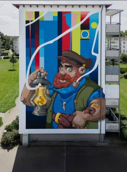 Streetartwall. An impressive mural (15.15 meters high and 9.5 meters wide) of a captain holding up a lantern was sprayed/painted on the outside wall of a multi-storey tower block. It unites two artists with different styles. The lively comic style by Hombre Suk with clear graphic elements by "The Cut" (Peter Stöcker). At the center is a maritime figure - a bearded, tattooed captain with a pipe, holding up an old lantern. The background is formed by colored geometric surfaces, small circles and white arcs that begin in the pipe and extend across the surface to the lantern.
Info: Peter Stöcker, Bremen artist and owner of the urban art agency "Lucky Walls", has realized an impressive art project with several well-known street artists under the motto "Meet each other". The design of the mural by "Hombre Suk" focuses on the power of encounters and exchange, both on a creative and social level. The mural is to be understood as an appeal to approach the unknown with courage and openness.