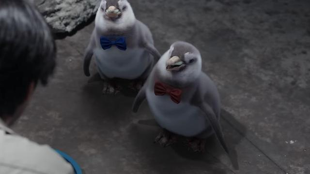 Two CG adolescent penguins wearing bow ties in blue and red stand on a rocky surface, facing a person.

One of the giveaways that make them look “off” is the sort of blurry rendering of their dense coat of feathers, an effect that makes them cartoonish. Also their bows just kind of float flush against their chests like they’re held in place by magic. 

Still adorable though.