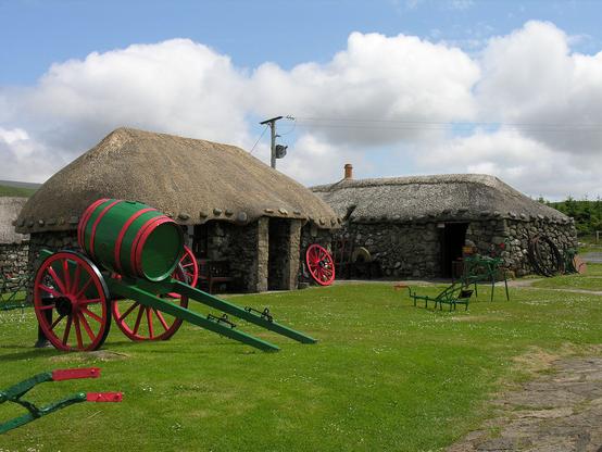 The Skye Museum of Island Life. The image shows two thatched stone single-storey buildings with a green and red cart in the left foreground that has a large barrel mounted on it. Other parts of green or red farming implements are visible bottom left and in the right middle of the frame. The scene is in sunlight.