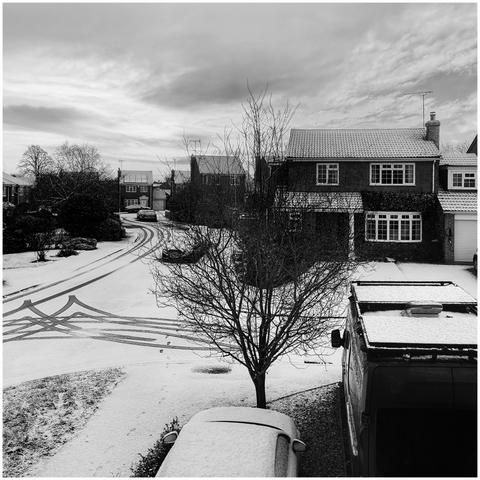 Monochrome shot of a winter morning. A fresh dusting of snow covers everything. Some dark tracks on the road show where a car has been. A show vibes car and van are in the foreground.