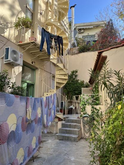 A charming little courtyard on two levels; humble, utilitarian, and pretty in a way that’s typical of vernacular architecture. On the left is a three storey building, of the simplest neoclassical style, painted yellow umber, with a green wooden door. There are concrete steps leading up to an upper courtyard, smaller and squarer, right in the middle. From the left side of this rises a wrought iron spiral fire escape staircase, also painted an earthy dusty yellow, with a landing on the storey above, which continues past the frame of the image. On the right is a little outbuilding with a ceramic tile roof, and behind, uphill, the top of a similar house and courtyard or garden. There are several pots of thriving plants all about, enhancing the space with living green, and brightly patterned sheets across a washing line on the lower level, jeans and leggings on another line at the fire escape landing. But for these and the air conditioning unit on the wall, it might be 100 years ago.