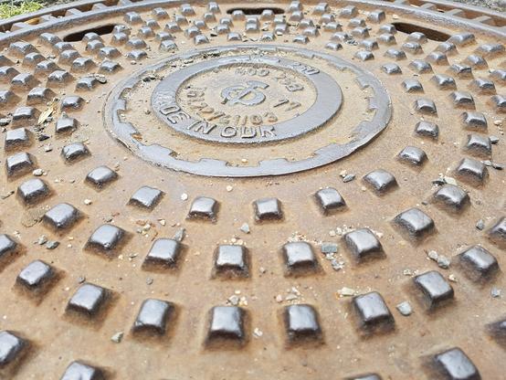 Close-up photo of a manhole cover showing its textured surface with concentric circles of raised square studs for traction. The center features a circular embossed design with text that reads "Made in GDR". The cover is made of metal with a brownish-gray coloring and shows some dirt and debris on its surface.