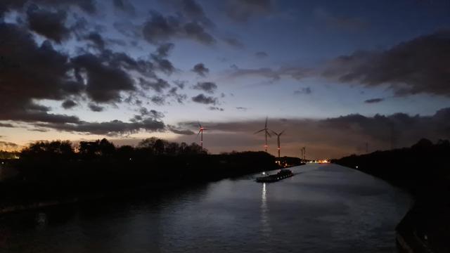 We are on a bridge. A wide canal going off to the horizon, some wind generators scattered in the background. The sky follows a gradient from a cold yellow through ice blue to deep blue, covered by dark clouds in the twilight. A heavily loaded barge appears as a shadow on the reflective gray water.