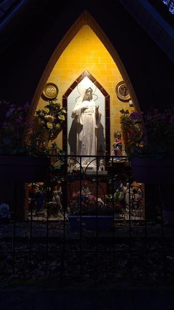 A statue of Maria outside in the light, at night. Surrounded by flowers and smaller religious statues. She's covered by a roof shaped like two arches, the wall behind her is lit and tiled. Yellow, black and white tiles.