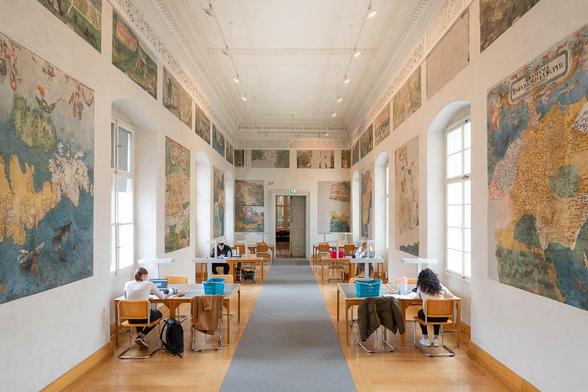 Interior of a library inside a Renaissance palazzo, showing a long and narrow reading room with work tables lining both walls. Between large vaulted windows, the walls are decorated with frescoes depicting maps.