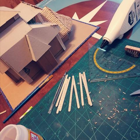 Foto von meiner Arbeitsplatte. Man sieht das Modell meiner norwegischen Stabkirche, hauptsächlich aus Pappe mit einigen verklebten Schindeln geschnitzt aus Kaffeerührstäbchen aus Holz. Dann meine Bastelmatte, ein Schnitzmesser, Holzleim, die Rührstäbchen und ein Geodreieck.