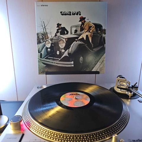 A vinyl record sits on a turntable. Behind the turntable, a vinyl album outer sleeve is displayed. The front cover shows Willie Colon and Hector Lavoe dressed in trench coats and hats around a classic car to give off mob vibes.