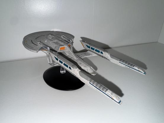 A detailed model of a futuristic spaceship displayed on a black stand against a white background. The ship features a sleek design with a circular saucer section and elongated wings, showcasing intricate details and markings.