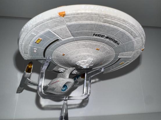 A detailed model of a starship, resembling the USS Titan from the Star Trek franchise, displayed from below. The model is mounted on a transparent stand against a plain background.