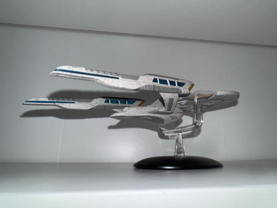 A detailed model of a sci-fi spaceship displayed on a stand, featuring a sleek design with blue and gray accents. The background is a neutral, light-colored surface.