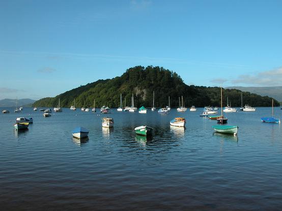 The east side of Loch Lomond at Balmaha and the island of Inchcailloch. The image shows the still blue waters of the loch extending into the frame past lots of small, moored boats that are all pointing towards us. The island stretches across the centre of the frame and is covered in trees. In the distance is the far side of the loch. The sky is mainly blue.