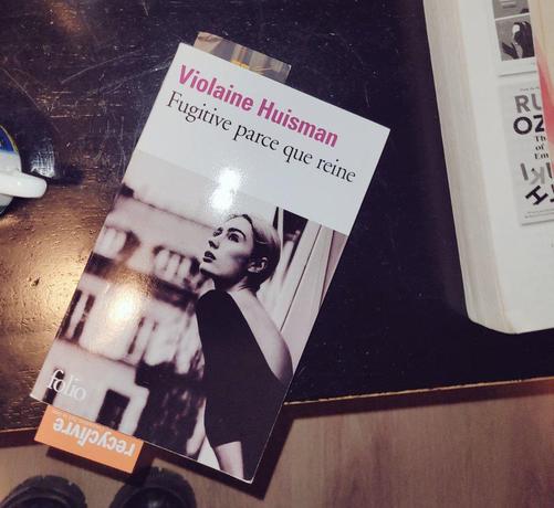 Un livre de poche posé sur une table basse, aux éditions Folio : première de couverture visible du livre "Fugitine parce que reine" de l'autrice Violaine Huisman (écrit en rose), avec une photo noir et blanc moderne d'une femme maquillée et les cheveux châtains attachés tendue à la fenêtre d'un appartement au dernier étage d'un bâtiment, comme ses voisins, de style Hausmanien, à Paris.