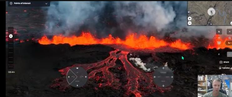 Screenshot of drone video from current Iceland eruption. The background is dominated by lava fountains stretching across the frame. A large tongue of lava is flowing toward the camera from a point in the center of the image.