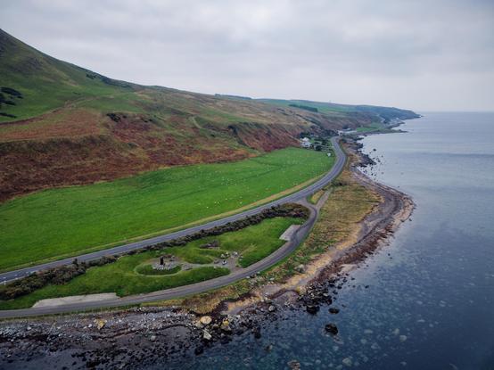 Drone photo from beside the memorial monument to the Varyag cruiser that ran aground offshore near Lendalfoot in the early C20. Looking inland, a flat plain makes a stark contrast with the subsequent elevated former coast / line of a raised beach.